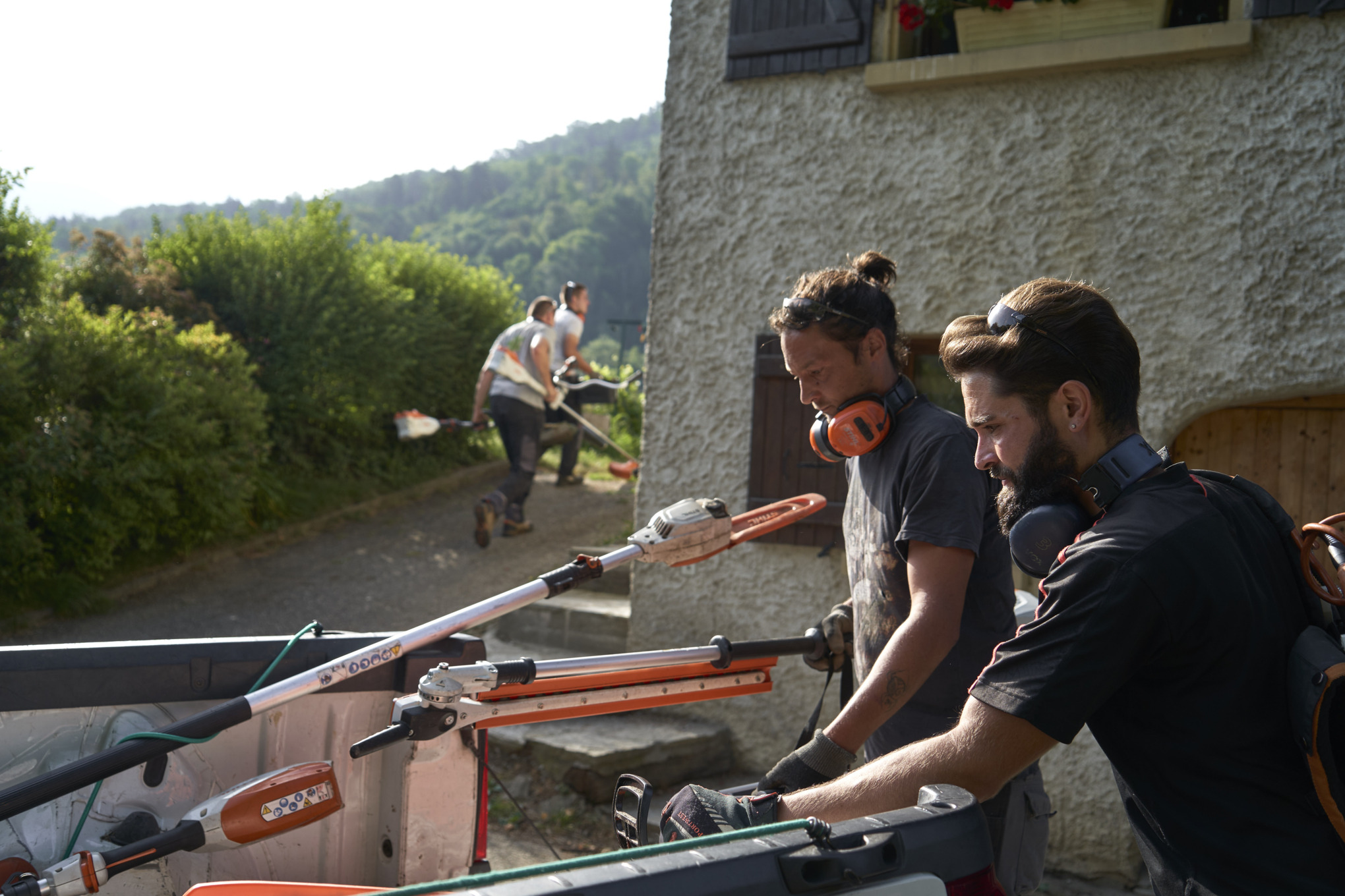 Two landscape gardeners unload STIHL cordless power tools from a car, with other workers in the background 