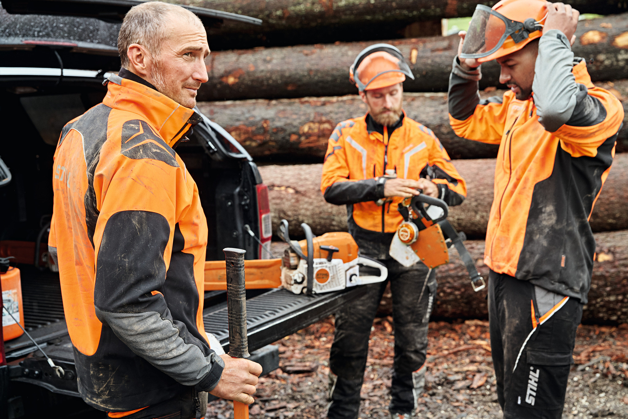 Three men in protective gear in front of a vehicle with an open trunk, STIHL cordless tools inside.