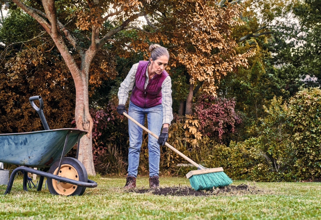 A woman sweeps leaves in a garden beside a metal wheelbarrow.