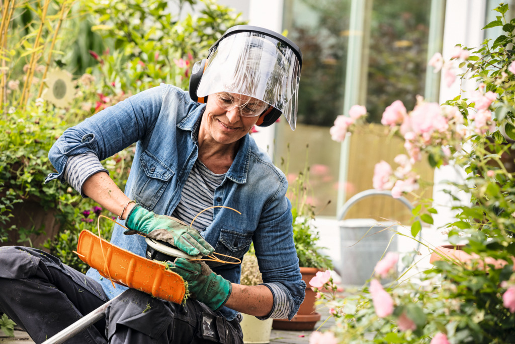 A woman wearing STIHL protective equipment walks along a path bordered by square-trimmed bushes with a cordless power tool in her hand