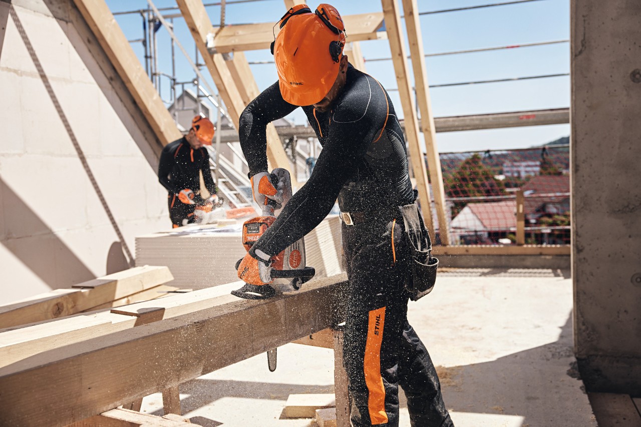 A STIHL professional under a roof structure saws a wooden beam with a battery chainsaw, while another worker uses a battery chainsaw in the background